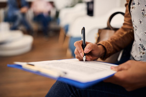 Woman’s hands filling out forms on blue clipboard in waiting room
